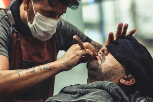 A masked barber in an apron shaves a client’s beard with a straight razor as the client reclines with a dark towel over his eyes